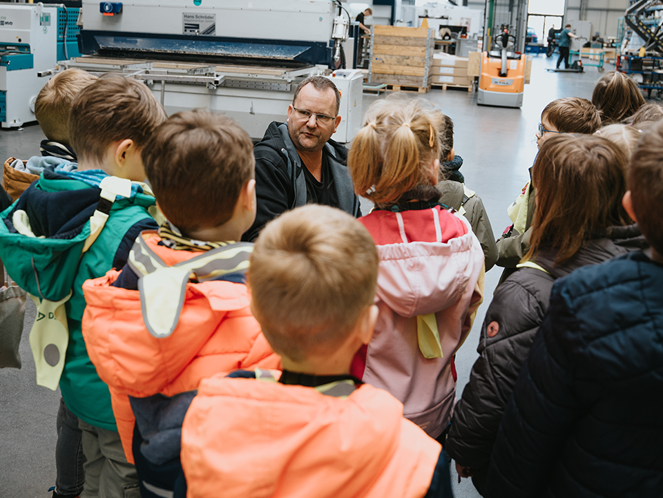 Rembe1 Kinder der Kita-Gruppe mit einem Mitarbeiter von Rembe in einer Produktionshalle. Die Kinder stehen mit dem Rücken zur Kamera. Der Mitarbeiter hockt vor den Kindern und sieht sie an.
