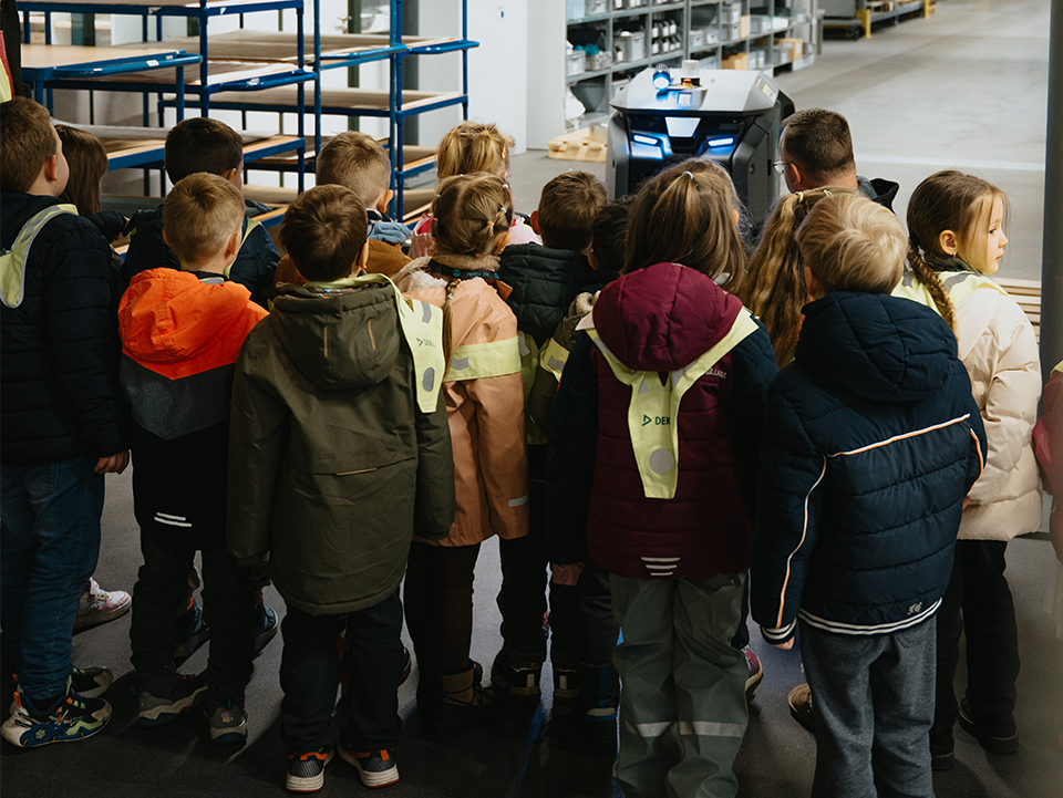Rembe 1 Eine Gruppe Kinder im Vorschulalter steht mit dem Rücken zur Kamera und blickt in eine Produktionshalle.
