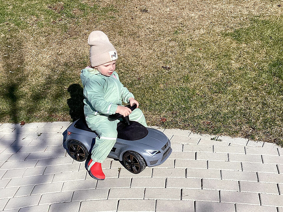 Ein Kind im Matschanzug und mit Mütze sitzt auf dem neuen Rutschauto. 