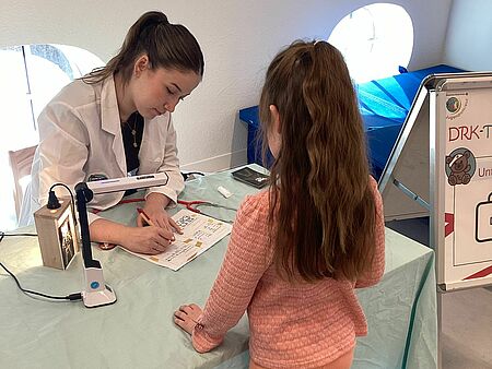 Die Teddyärztin sitzt an einem Tisch, davor steht ein Mädchen im Vorschulalter mit langen dunklen Haaren. Die junge Frau, die die Ärztin ist, trägt den Befund des Kuscheltieres in die Akte ein. Auf dem Tisch steht eine sehr helle Lampe, die als Röntgengerät für die Kuscheltiere genutzt wird. 