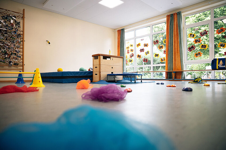 In der Turnhalle ist eine Bewegungslandschaft aufgebaut. Im Zentrum steht ein großer Kasten mit einem kleinen Trampolin davor und einer Matte dahinter. Im Vordergrund liegen Tücher auf dem Boden. Am linken Bildrand sind kleine Hürden mit gelben und blauen Hütchen aufgebaut. Am rechten Bildrand ist eine große Fensterfront, davor steht eine Bank. Außerdem liegen Säckchen und andere Utensilien auf dem Boden.
