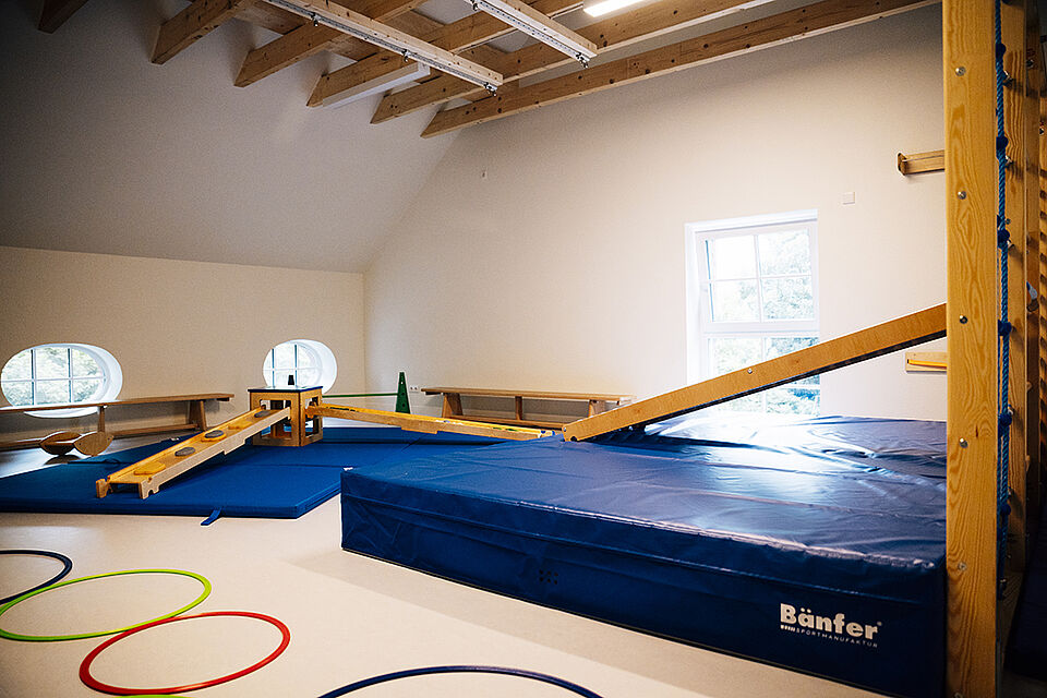 In der Turnhalle ist eine Bewegungslandschaft aufgebaut. Es ist seitlich eine Sprossenwand zu erkennen, davor liegt eine große Turnmatte und eine Leiter ist in die Sprossenwand eingehängt. Im Vordergrund liegen Reifen auf dem Boden.