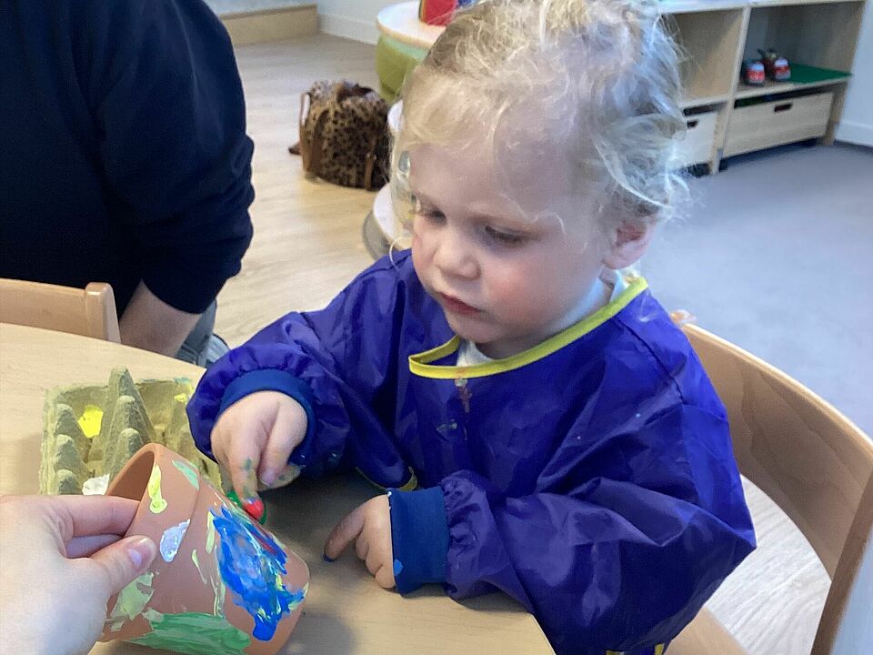 Ein 2 bis 3-jähriges Kind in einem blauen Malkittel betupft einen kleinen Tontopf mit Farbe. Dabei sitzt es auf einem Stuhl an einem Tisch. Der Tontopf wird von einer Hand festgehalten.