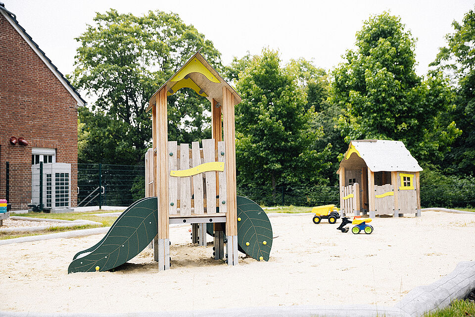 Der Spielbereich für die kleineren Kinder mit einem kleinen Kletter- und Spielelement aus Holz. Es hat eine grüne Rutsche auf der einen Seite und eine grüne Treppe auf der anderen. Im Hintergrund ist ein kleines Holzspielhaus zu sehen. Beide Elemente stehen im Sand, auf dem noch ein Bagger und anderes buntes Sandspielzeug liegt.