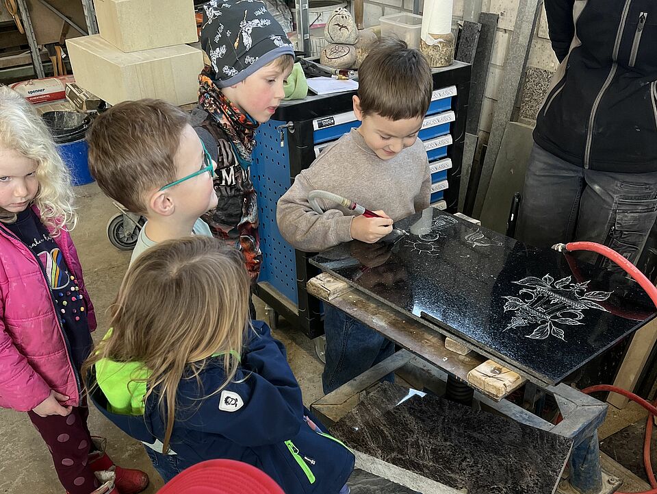 Die Kinder stehen um eine schwarze Steinplatte herum in der Werkstatt und können sie mit einer kleinen Fräse selbstständig bearbeiten.