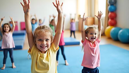 Eine Gruppe Kinder steht auf einer großen Turnmatte und heben die Arme in die Höhe. Die Kinder sind bunt und sportlich gekleidet. Im Hintergrund sieht man große Bölleund weitere Turnmatten.