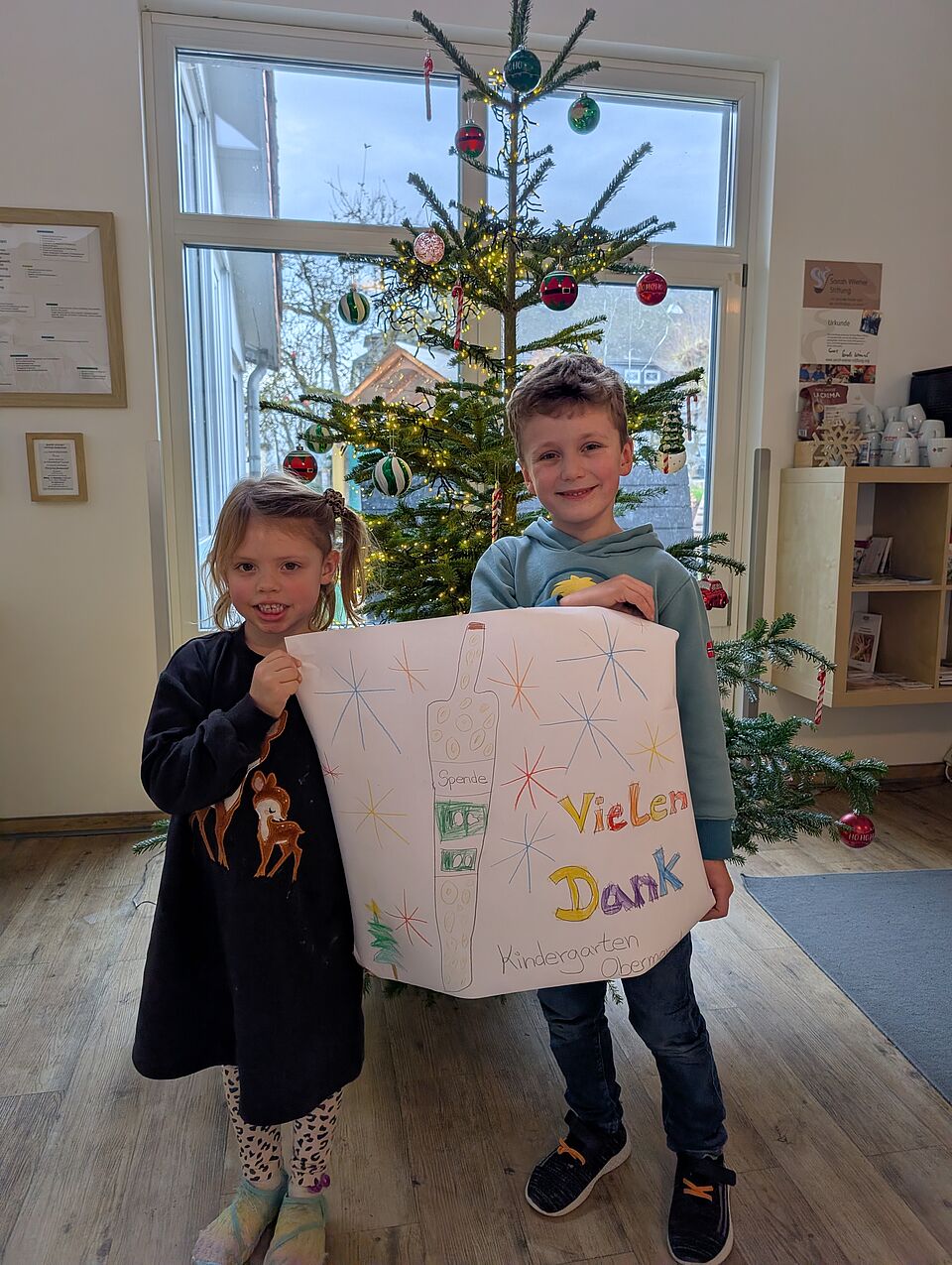 Ein Mädchen und ein Junge stehen vor einem geschmückten Weinachtsbaum und halten ein gemaltes Bild in der Hand. Auf dem Bild sind Sterne und die Spendenflasche aufgemalt. Dabeben steht "Vielen Dank" und "Kindergarten Obermarsberg".