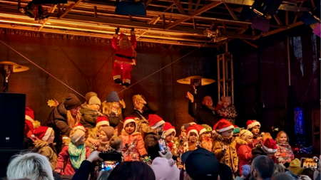 Kinder stehen vor der Bühne eines Weihnachtsmarktes. Auf der Bühne hängt ein Weihnachtsmann von der Decke.