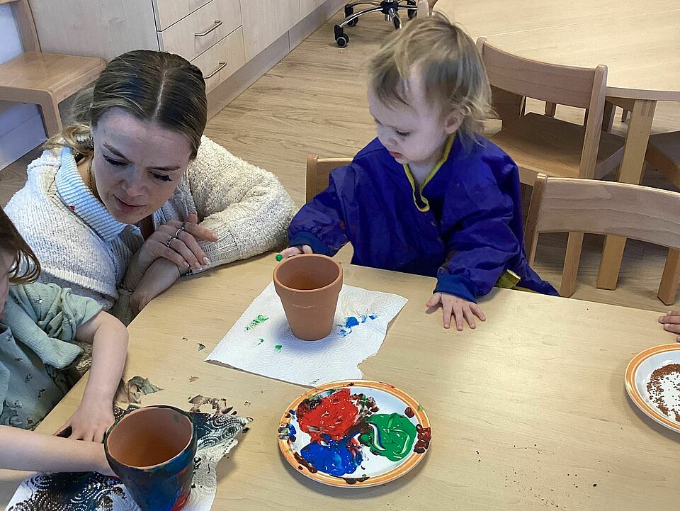 An einem Tisch sitzen zwei Kleinkinder, zwischen ihnen hockt eine Frau am Tisch. Die Kinder geben Erde in die kleinen Tontöpfe.