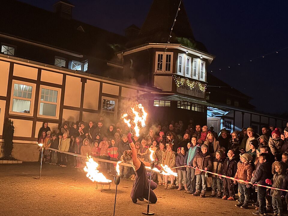 IMG 8416 Ein Feuerkünstler, der Tricks vor einem großen Publikum vor der Schützenhalle zeigt. Der Künstler hat drei Stangen die er wild umherwirft in der Hand. Die Stangen sind im Brennen
