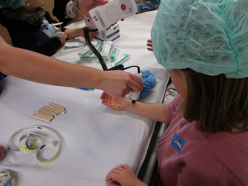 Bei einem Mädchen wird der Puls gemessen am Handgelenk gemessen von einem Mitarbeiter des Krankenhauses. Das Mädchen hat eine Haube für den OP auf dem Kopf. 