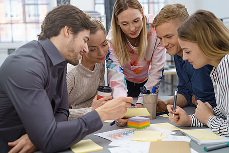 Zwei Männer und zwei Frauen rund um die 30 unterhalten sich angeregt. Vier sitzen an einem Tsich mit Schreibmaterialien, eine Frau steht vorgebeugt am Kopf des Tisches. Alle schauen interessiert auf den Tisch.