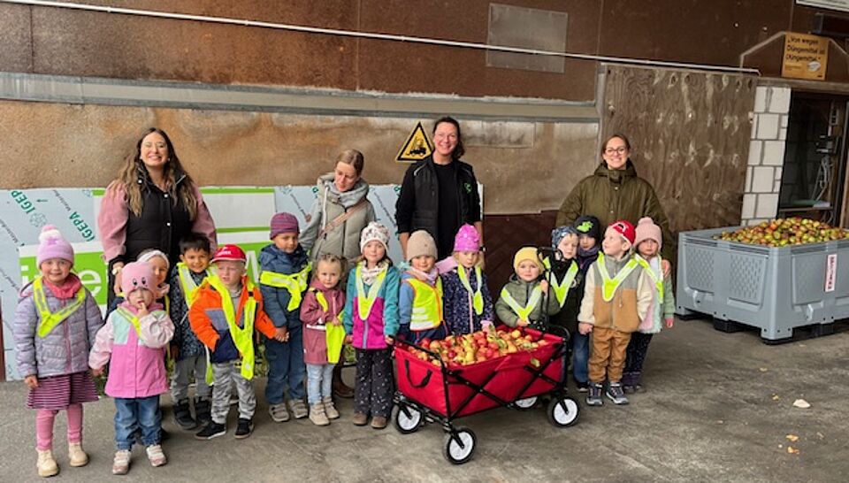 Kinder der Kita des DRK Familienzentrums Brilon stehen mit ihren Erzieherinnen und einem Bollerwagen voller Äpfel bei der Raiffeisen Genossenschaft.