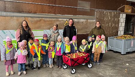 Kinder der Kita des DRK Familienzentrums Brilon stehen mit ihren Erzieherinnen und einem Bollerwagen voller Äpfel bei der Raiffeisen Genossenschaft.