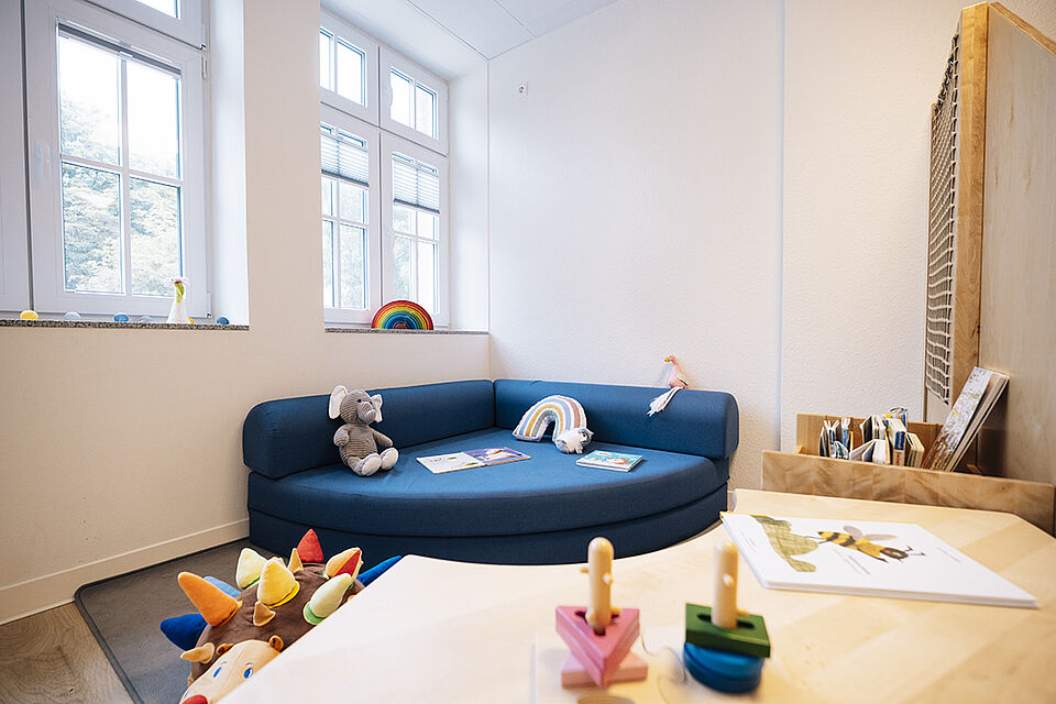 In der Ecke des hellen Raums steht ein großes, blaues Ecksofa, auf dem Bücher liegen. Davor liegt ein großer Plüschigel und im Vordergrund ist ein Tisch zu erkennen mit einem Stapelspiel darauf.