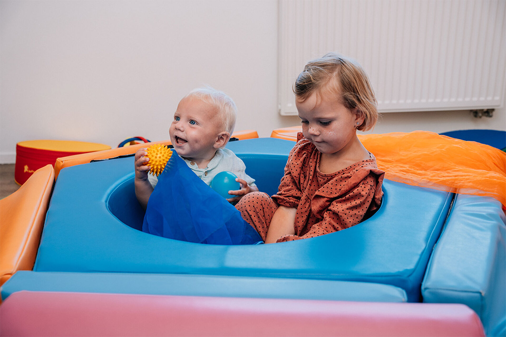 Zwei kleine Kinder sitzen auf weichen, bunten Schaumstoffmatten. Das Kind links hält ein gelbes Spielzeug in der Hand und schaut nach links. Es lacht. Das Kind rechts trägt ein rotes Kleid mit schwarzen Punkten und schaut nach unten. Die Matten sind blau, orange, pink und rot. Im Hintergrund ist eine weiße Wand mit einer Heizung zu sehen.