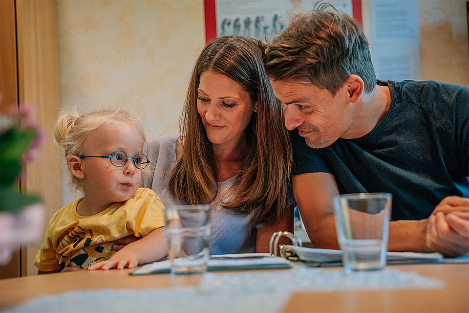 Eine junge Familie, die Eltern sind circa Anfang bis Mitte dreißig und ein kleines Mädchen mit ungefähr zwei Jahren sitzen an einem Tisch. Das Mädchen links sitzt auf dem Schoß der Mama, der Papa sitzt rechts daneben. Die Eltern schauen ihr Kind erwartungsvoll an. Das Kind hat blonde seitliche Zöpfe und trägt eine Brille. Alle sind leger gekleidet. Es stehen zwei Wassergläser auf dem Tisch und es sind Unterlagen zu erkennen.