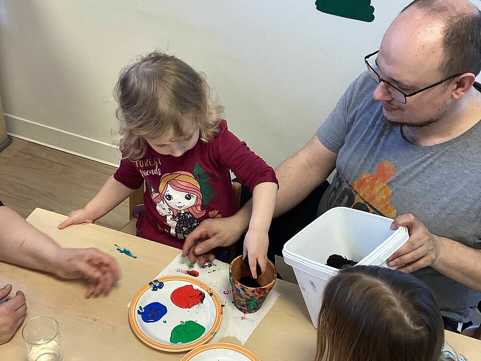 Vater und Tochter sitzen nebeneinander am Tisch und streuen die Kressesamen auf die Erde im Tontopf.