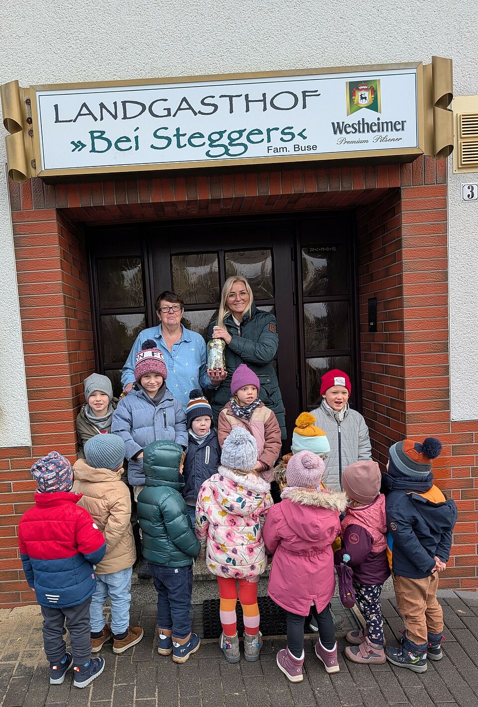 Die Wirtin des Landgasthofs, Marianne Buse, und Britta Gerlach, Stadortleitung DRK KiTa Obermarsberg, stehen mit 12 Kindern der Kita im Eingang vor dem Gasthof. Alle sind winterlich gekleidet. Die vorderen sieben Kinder stehen mit dem Rücken zu Kamera, die anderen fünf und hinten die beiden erwachsenen Frauen gucken in die Kamera. Britta Gerlach hält das Spendenglas mit dem Geld in der Hand.  