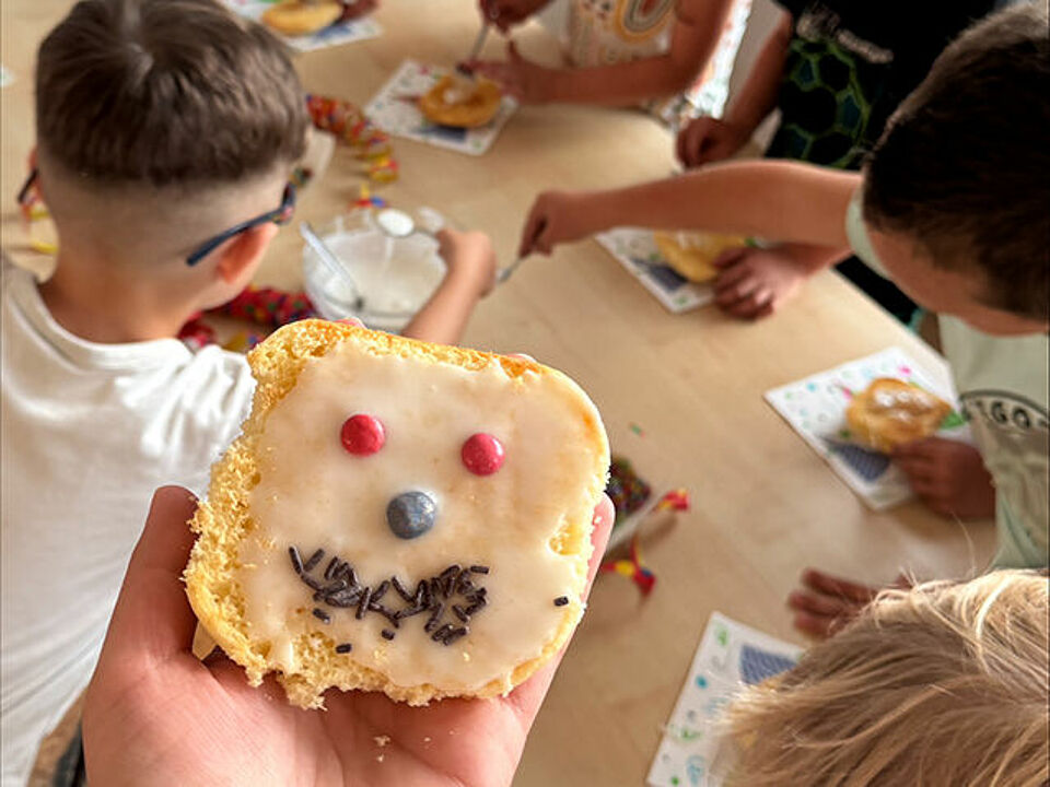 Die Kinder verzieren Amerikaner, ein Amerikaner mit einem Gesicht aus Streuseln und Smarties wird in die Kamera gehalten.
