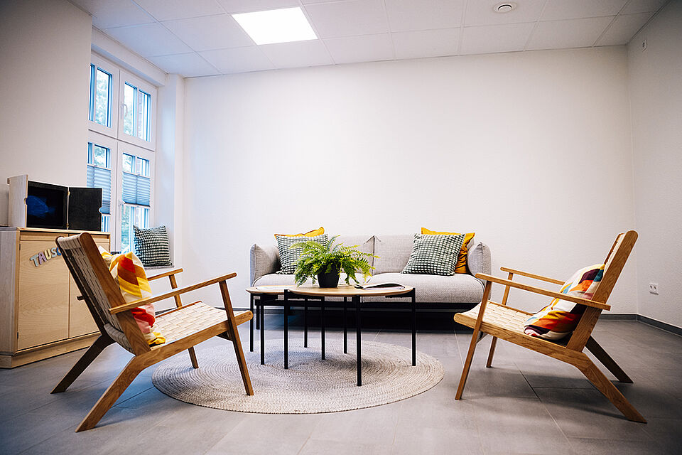 In einem hellen Raum stehen zwei Relaxstühle und ein kleines Sofa, in der Mitte steht ein niedriger zweiteiliger Tisch und auf dem Boden liegt ein Teppich. Auf dem Tisch stehen Blumen.
