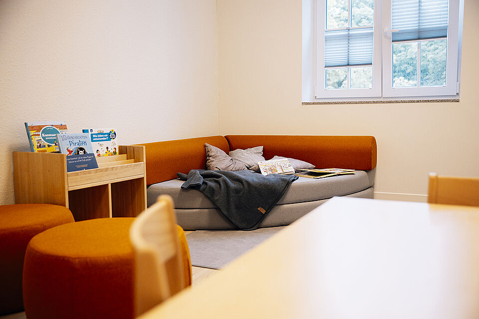Im Nebenraum eines Gruppenraums steht ein großes orangebraunes Ecksofa. Auf dem Sofa liegen Decken, Kissen und Bücher. Daneben steht ein Bücherregal und im Vordergrund noch Hocker, die zum Sofa passen.