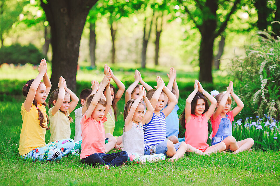 Eine Gruppe von ungefähr 10 Kindern sind bunt gekleidet und sitzen im Gras. Sie sitzen im Schneidersitz und verschließen die Hände über den Köpfen.