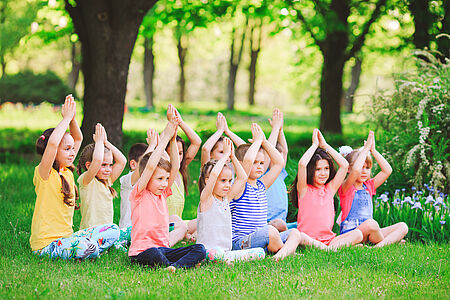 Eine Gruppe von ungefähr 10 Kindern sind bunt gekleidet und sitzen im Gras. Sie sitzen im Schneidersitz und verschließen die Hände über den Köpfen.