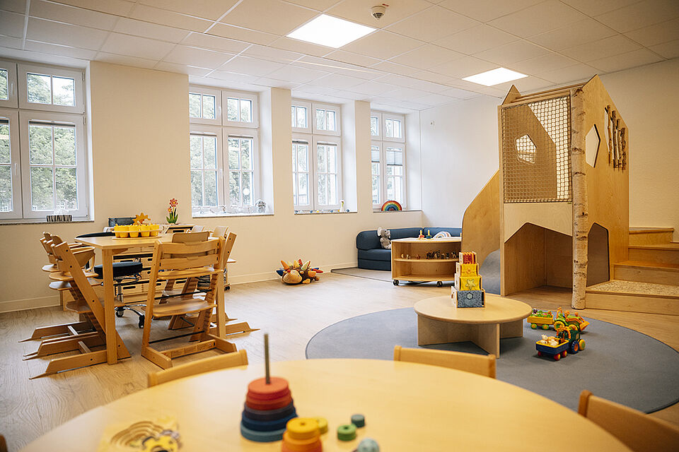 Ansicht eines Gruppenraums für die kleinen Kinder. Es ist ein heller mit Raum mit einem Tisch, an dem Hochstühle stehen und einem Spielelement, dass den Kindern ermöglicht, zu klettern. Außerdem gibt es noch kleine Tische mit altersgerechtem Spielzeug darauf.
