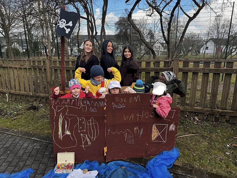 Mittig auf dem Bild sieht man ein selbstgebautes und verziertes Piratenschiff, hinter dem sechs Kinder zwischen 4 und 6 Jahren stehen. Hinter ihnen stehen drei junge Frauen um die 20. Alle gucken in die Kamera und präsentieren ihr gebautes Werk im Garten der Kita.. 