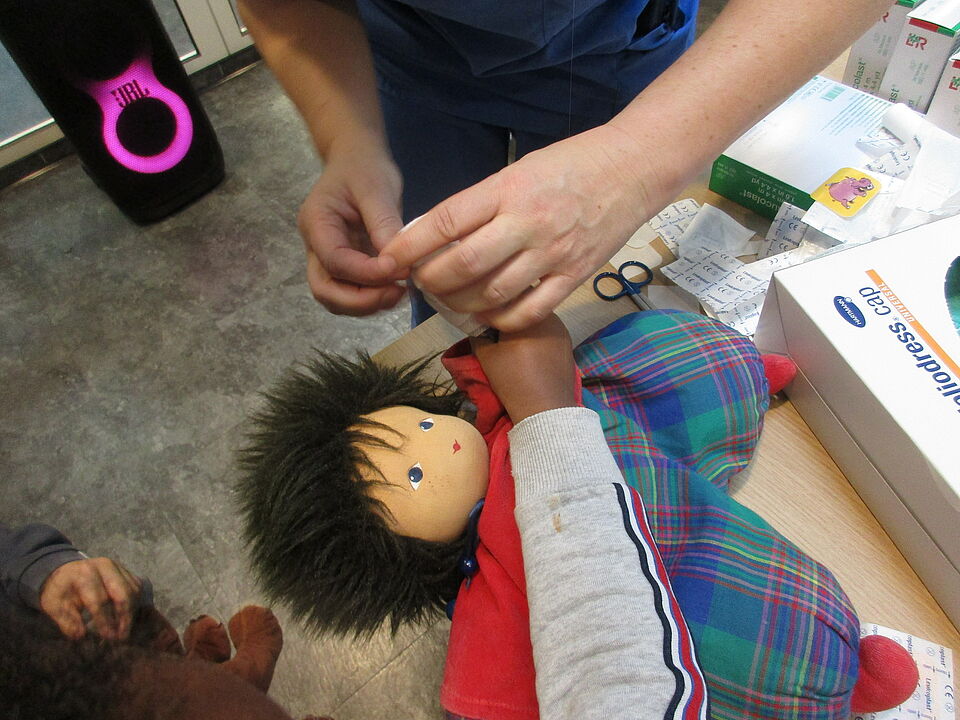 Die Puppe Maxi wird in der Teddyklink verarztet. Man sieht die Puppe, die auf einem Tisch liegt und von einen Verband bekommt.  