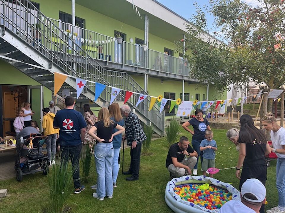 Im Garten der KiTa steht eine Gruppe Eltern mit einer Erzieherin am Planschbecken und schauen zu, wie jemand versucht, Bälle darauf zu fischen. Andere Erwachsene stehen in einer Gruppe vor dem Tisch, an dem die Kinder geschminkt werden.