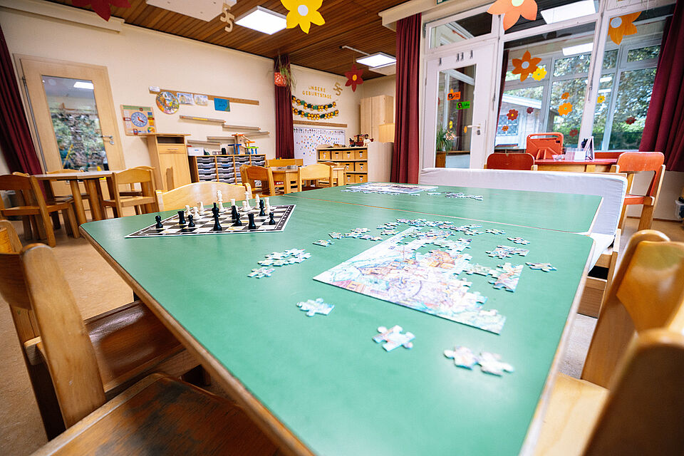Im Vordergrund steht ein großer Tisch mit einer grünen Tischplatte. Darauf liegt ein Puzzle, dass noch nicht fertig ist. Links daneben ist ein Schachspiel aufgebaut. Im Hintergrund ist der Gruppenraum zu sehen.