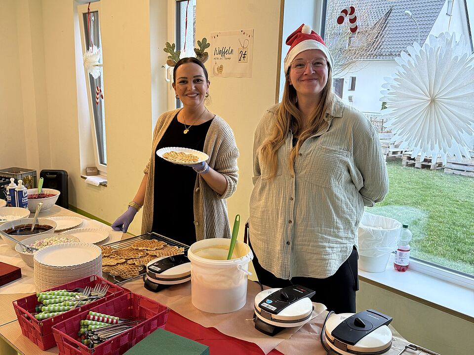 6867 Zwei Erzieherinnen stehen hinter einem Tisch mit Waffeln und lächeln in die Kamera und präsentieren eine Waffel