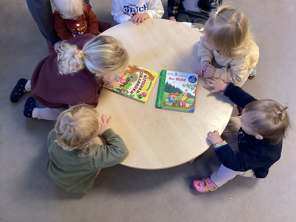 Sieben Kleinkinder sitzen um einen Kindertisch. Sie schauen auf zwei Bücher, die auf dem Tisch liegen. Eines hat einen ukrainischen Titel und ist in kyrillischer Schrift geschrieben. Das andere Buch ist eins aus der Reihe Wieso? Weshalb? Warum? Junior und heiißt "Der Wald".