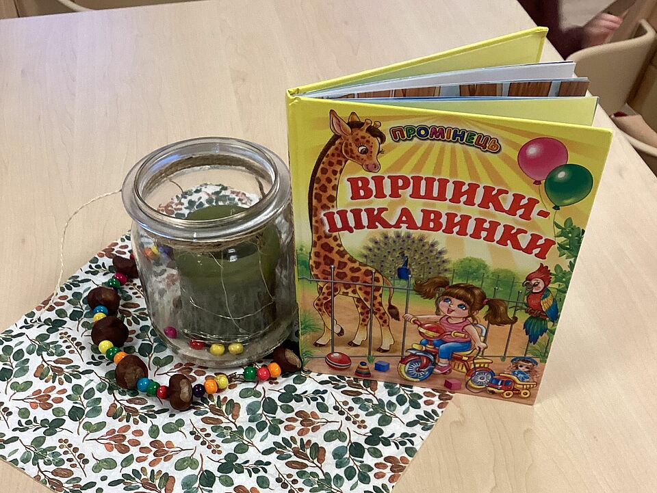Auf einem Tisch liegt eine herbstlich gestaltete Papierserviette, auf der ein Windlicht steht und daneben steht ein Kinderbuch mit ukrainischemText. Auf dem Titel ist eine Giraffe, ein Pfau, ein Papagei und ein Mädchen mit dicken braunen Zöpfen auf einem Dreirad zu sehen. Das Mädchen und die Giraffe schauen sich freundlich und neugierig an. 
