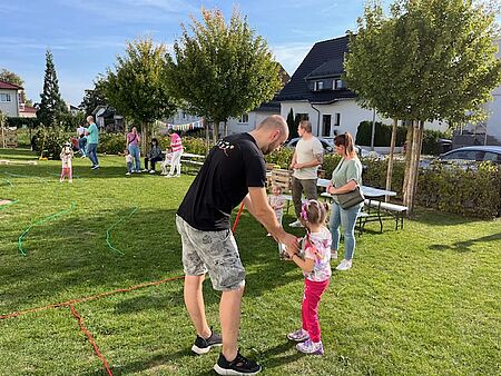 Ein Erzieher und ein Mädchen stehen im Vordergrund, mehrere Erwachsene stehen dahinter und schauen auf ein Mädchen, das im Hintergrund steht und ein Steckenpferd in der Hand hat.