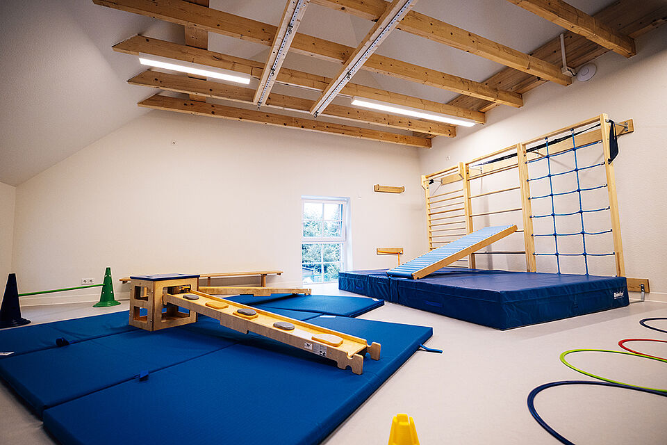Im Vordergrund liegen zwei große blaue Turnmatte, auf denen Bänke und Kästen zum Balancieren stehen. Im Hintergrund ist die Sprossenwand mit Leiter und Turnmatte zu sehen. Unter der Decke sind Holzbalken, an denen Haken hängen, um dort noch andere Dinge befestigen zu können.