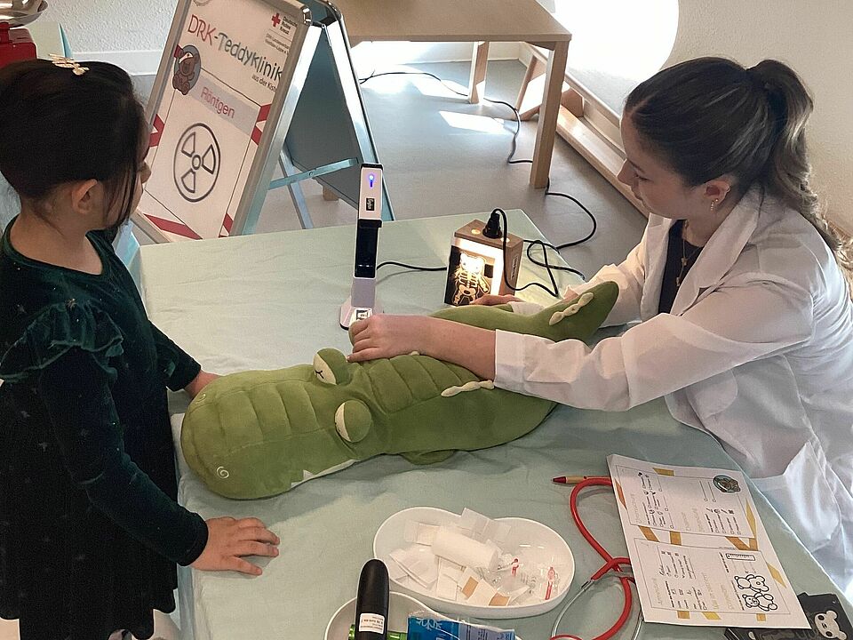 Ein großes grünes Stoffkrokodil wird von der Ärztin untersucht. Ein ca. sechsjähriges Mädchen schaut sich die Untersuchung genau an. Auf dem Tisch vor der Ärztin liegt eine Nierenschale, ein Stethoskop und die Akte des Kuscheltieres. 