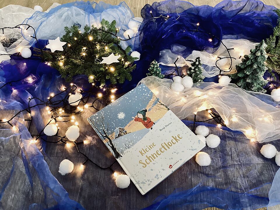 Das Kinderbuch "Die Schneeflocke" liegt auf transprarenten Tüchern in Blautönen. Auf den Tüchern liegen auch Lichterketten, Schneeflocken, Tannengrün und kleine Tannenbäume aus Keramik stehen als Deko mittendrin. 