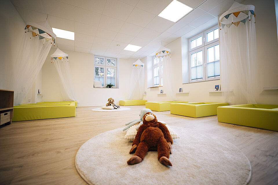 Ein großer Raum, in dem mehrere Schaumstoffbetten auf dem Boden stehen. Ein großer runder Flauschteppich liegt im Vordergrund, auf dem ein Kuschelaffe liegt. An der Decke hängen über den Betten kleine Baldachine mit transparentem Netzstoff.