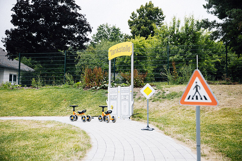 Es ist die Bobbycar-Rennstrecke zu sehen. Im Vordergrund steht ein Straßenschild "Achtung Fußgängerüberweg", im Hintergrund stehen zwei Rutschautos vor der Tankstelle.