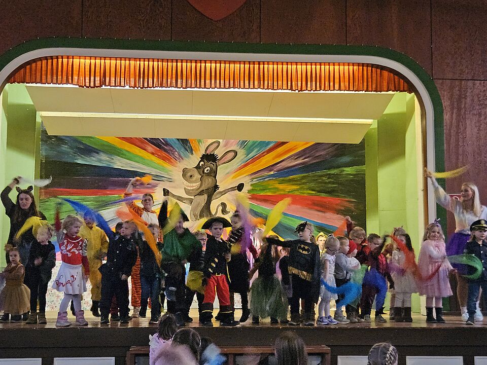 Verkleidete Kinder zwischen 3 und 6 Jahren stehen auf einer Bühne. Sie haben unterschiedliche Kostüme an und bewegen sich. Im Hintergrund der Bühne ist ein buntes gemaltes Bild in Regenbogenfarben. In der Mitte ist ein Esel im Comicstil, der lacht und sehr fröhlich aussieht.