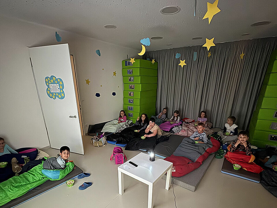 Die Kinder liegen im Schlafraum der Kita auf ihren Matten und in ihren Schlafsäcken und schauen in die Kamera
