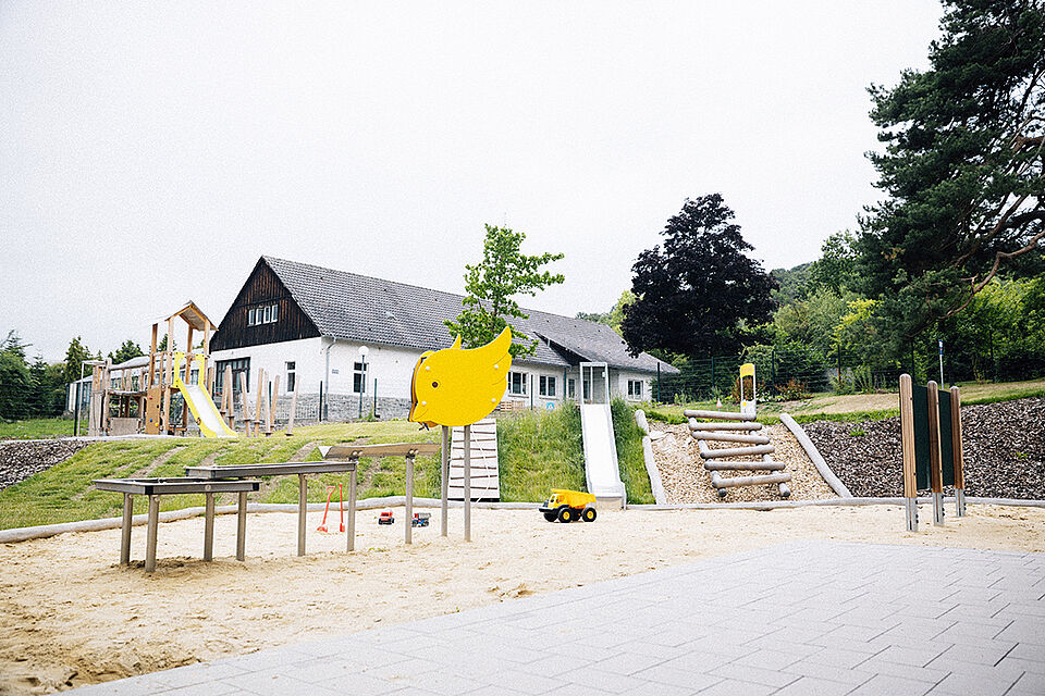 Blick vom Ausgang in den Garten auf das Außengelände. Links im Sand befindet sich eine große Matschanlage, dahinter sind in den Hang eine Rutsche und Klettermöglichkeiten eingebaut.