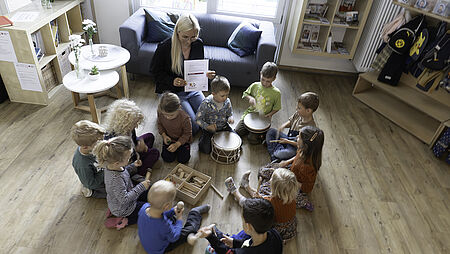 Kinder sitzen mit der Erzieherin auf dem Boden im Kreis und machen Musik.