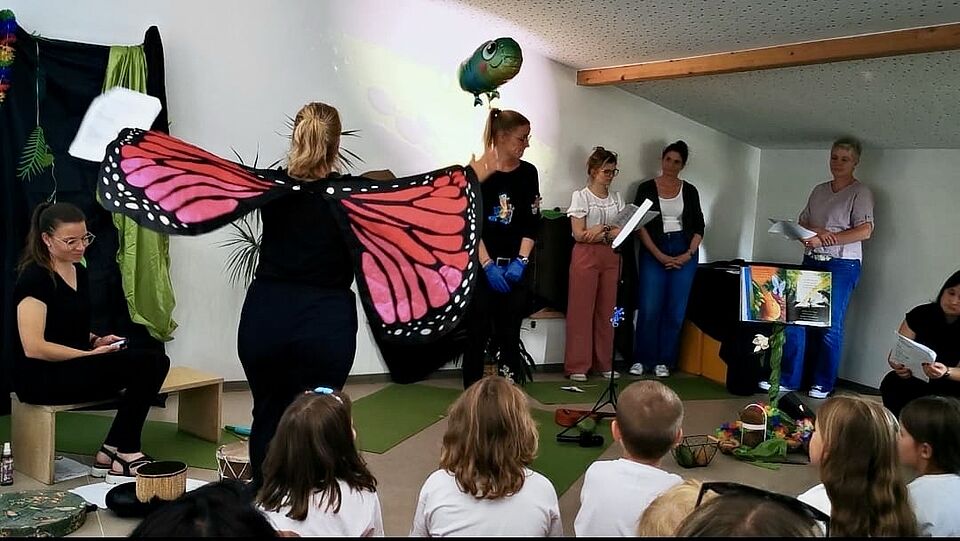 Die Erziehrinen führen vor den Kindern ein Theaterstück auf, alle sind schwarz angezogen, eine Person ist als Schmetterling verkleidet.