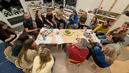 Herr Heinze zusammen mit den Eltern sitzen in einem Kreis am Elternabend. Er erzählt ihnen etwas und die Eltern hören aufmerksam zu.