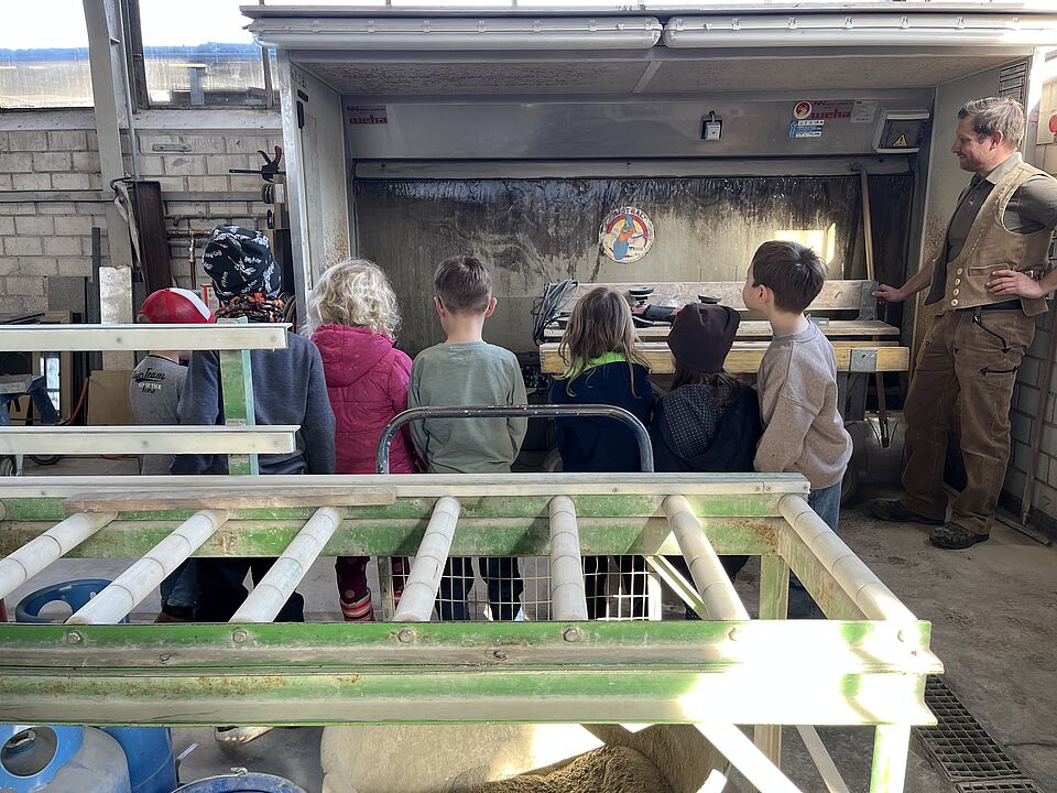 Der Steinmetz steht in Handwerkerkluft recht im Bild an einer Maschine und zeigt den Kindern, die mit dem Rücken zur Kamera stehen und auf die Maschine schauen, einen weiteren Arbeitsvorgang. 