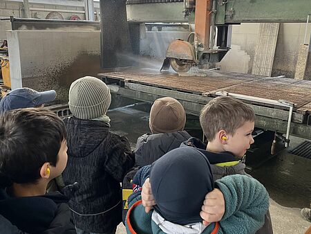 Die Kinder stehen vor einer Steinsäge, die eine Steinplatte schneidet. Wasser spritzt von der Stelle, an der gesägt wird, bis dan die ca. einen Meter entfernte Wand. Die Kinder stehen in einer Gruppe davor, sie tragen Jacken und haben Ohrstöpsel im Ohr. 